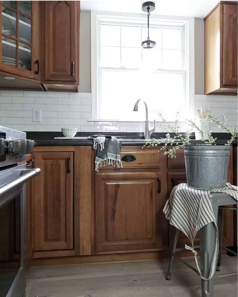 White subway tile backsplash kitchen is equipped with wood cabinets and black countertops. Black and glass pendant lighting hangs in front of kitchen window over single handle faucet. A rustic metal bucket rests upon a matching stool. Oak Cabinets White Quartz Countertops, Brown Kitchen Black Countertop, Dark Wood Kitchen Cabinets Black Countertop, Backsplash With Black Countertops And Wood Cabinets, Black Countertop Backsplash Kitchen, Oak Cabinets With Black Countertops And White Backsplash, Backsplash For Black Granite Countertops And Brown Cabinets, Brown Kitchen With Black Countertops, Dark Brown Cabinets Black Countertops