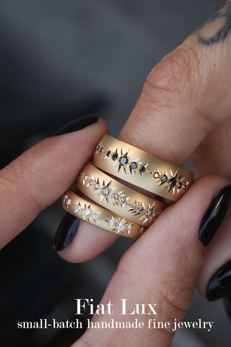 A light skinned hand holds three yellow gold rings of similar design in three sizes with white diamonds, champagne diamonds, and black diamonds. Each ring has repeating sets of diamonds surrounded by hand engraving that looks like a starburst. Maiden Voyage Jewelry, Alternative Wedding Bands For Women, Gender Neutral Wedding Bands, Gender Neutral Jewelry, Unisex Wedding Rings, Gender Neutral Wedding Ring, Unisex Engagement Rings, Gender Neutral Engagement Rings, Untraditional Wedding Rings