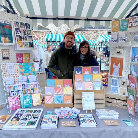 The photograph shows a man and a woman standing in thick winter layers and smiling, behind a market table stall with temporary walls attached at either side, to a green & white stripy gazebo. The products on display are brightly coloured cards, calendars, notebooks, prints, mugs and tote bags, with illustrations of dogs, cats, rabbits, goldfish, hamsters and more! Craft Fair Display Table, Booth Display Ideas Diy, Art Fair Display, Craft Fair Table, Market Stall Display, Christmas Market Stall, Art Fair Booth, Stall Display, Craft Market Display