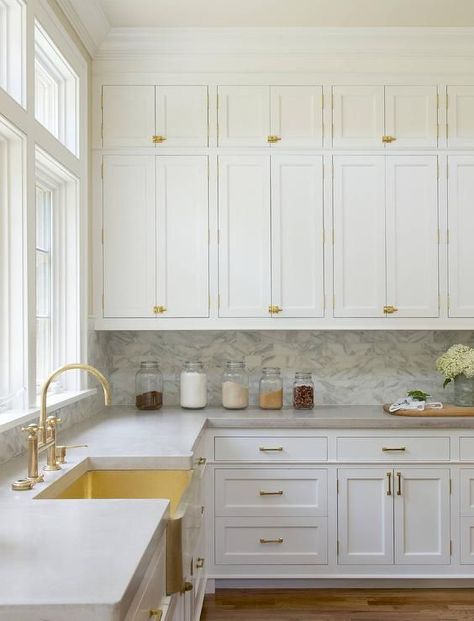 White stacked kitchen cabinets adorning vintage brass latch hardware are fixed above a marble backsplash lining white shaker cabinets topped with a gray quartzite countertop continuing around to a brass apron sink complemented with a brass gooseneck faucet positioned in front of a window. Kitchen With White Cabinets, Backsplash For White Cabinets, Kitchen New York, White Shaker Cabinets, Kitchen Cabinets Makeover, Classic Kitchen, Marble Counter, Kitchen Cabinet Hardware, Home Luxury