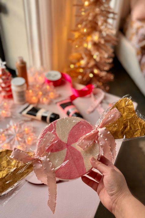 Oversized candy ornament finished craft is shown in the foreground while the background shows a pink desk with aesthetic DIY supplies like paint, glitter, paint brushes, and bows. The oversized candy is a pink peppermint painted on styrofoam wrapped in cellophane and using pink ribbon to tie the ends. Very whimsical Christmas vibe! Christmas Crafts Candy, Diy Pink Christmas Decorations, Plum Decor, Oversized Ornaments, Candy Ornaments, Door Decorating, Candy Theme, Styrofoam Ball, Pink Sugar