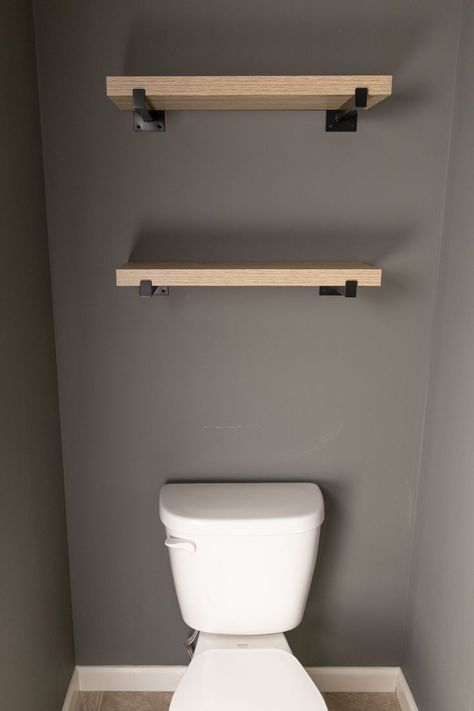 A water closet with dark gray walls, a white toilet and two floating shelves above the toilet. These shelves have two wicker baskets to organize feminine products and toilet paper. White Shelves With Black Brackets, Storage Above Toilet Ideas, Styling Floating Shelves, Shelves Behind Toilet, Bathroom Organizer Ideas, Shelf Above Toilet, Shelves Above The Toilet, Two Floating Shelves, Over The Toilet Organizer