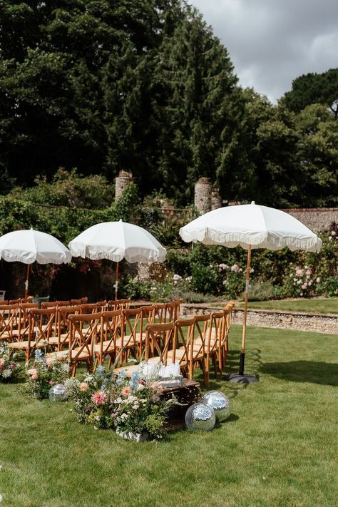 This shows a sunny outdoor wedding ceremony. The focus is on rows of wooden cross back chairs along the aisle. Next to each row of chairs are white parasols. At the other end of the row are colourful floral arrangements and disco balls. Bench Wedding Seating, Banquette Seating Wedding, Cross Back Chair, Dried Foliage, Trestle Tables, Wedding Ceremony Seating, Crossback Chairs, Banquette Seating, Wedding 2025