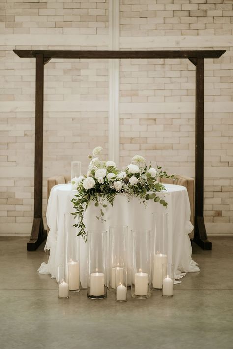 This sweetheart table is decorated with a lush floral arrangement featuring white roses, baby's breath, queen anne's lace, eucalyptus, and italian ruscus. Floral design by Imogen Floral Design. White Florals Sweetheart Table, Sweetheart Table White Flowers And Greenery, Wedding Sweetheart Table With Candles, Sweetheart Table Wedding Candles On Floor, Sweetheart Table Wedding Eucalyptus, Sweetheart Table With Eucalyptus, Head Table Wedding Decorations Candles, Minimalist Wedding Sweetheart Table, Sweetheart Table Wedding With Candles