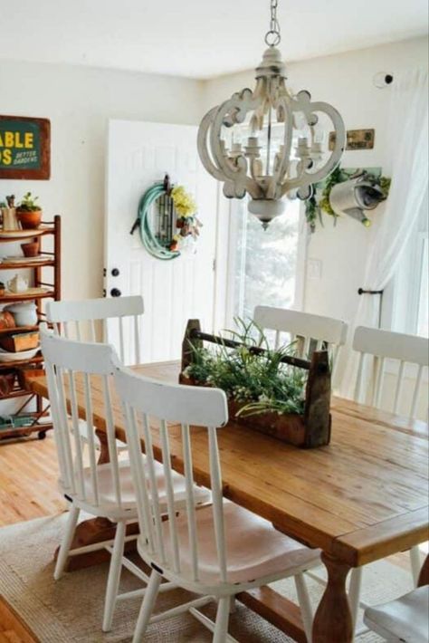 Step into a dining room brimming with garden-inspired beauty and vintage charm. A rustic warm wood dining table stands at the heart, encircled by white wooden chairs that rest on a textured area rug. An antique wooden toolbox, filled with verdant greenery, serves as a delightful centerpiece. Above, a vintage white chandelier illuminates the scene. On the opposite side, a warm wood shelf adorned with serving ware and potted greenery graces a white wall. White Dining Chairs Wood Table, Wooden Table White Chairs, White Wood Dining Chairs, White Wooden Chairs, Mismatched Chairs, Rustic Farmhouse Table, Stained Table, Vintage Inspired Decor, White Dining Chairs