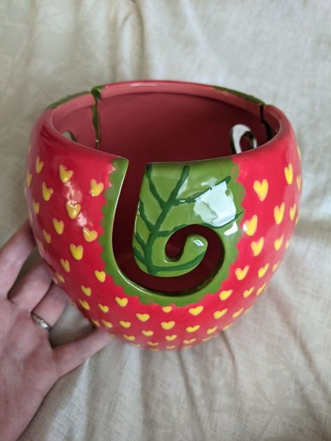 A red yarn bowl with yellow hearts painted all over it to look like a strawberry. The yarn bowl has a ceramic swirl yarn feeder that is painted green to mimic a leaf. A hand props up the bowl against a cream fabric backdrop. Ceramic Yarn Bowl Painting Ideas, Pottery Crochet Bowl, Cute Clay Yarn Bowl, Yarn Bowls Pottery Diy, Painted Yarn Bowl, Yarn Bowls Pottery Painting, Polymer Clay Yarn Bowl, Yarn Bowl Painting Ideas, Knitting Bowls Pottery