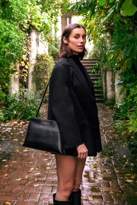 Tall brunette model with short brown hair wears a black coat, black shorts and high black boots. She stands on an abandoned pergola, with pillars covered in green ivy and leaves. She has a crossbody bag over her shoulder. The bag is made black grain textured vegan leather, which shimmers in the light. It's softly structured with two pouches joined together. It has a thin leather crossbody strap. Black Crossbody Bag Outfit, Lost Woods, Crossbody Bag Outfit, Purse Outfit, Book Wallet, Halter Swimwear, Vegan Handbags, Luxury Crossbody, Crossbody Bag Black