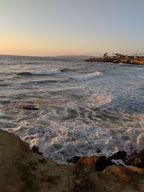 Image of the view from Sunset Cliffs in San Diego California. Image reveals a brown rocky floor in the bottom fourth, a beautiful blue and white ocean full of movement, and a few incoming new waves below. In the distance in the top right corner is a few beach houses and many palm trees that overlook the ocean. Time of day is sunset and the lighting is soft. The sky has a hint of yellow, pink, and orange on the horizon. Santa Monica Beach Aesthetic, Anaheim California Aesthetic, San Diego Astethic, Priscilla Core Aesthetic, San Diego Beach Aesthetic, San Diego California Aesthetic, San Diego Vibes, California Summer Aesthetic, Laguna Beach Aesthetic