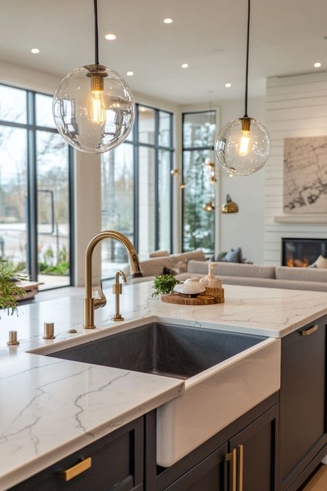 Modern kitchen with marble countertops, brass fixtures, hanging globe lights, and a view of a living area with a fireplace. Sink In Island Kitchen, Kitchen Sink Inspiration, Barn Sink, Apron Sinks, Kitchen Sink Ideas, Kitchen Sink Decor, Undermount Stainless Steel Sink, Modern Kitchen Sinks, Sink In Island