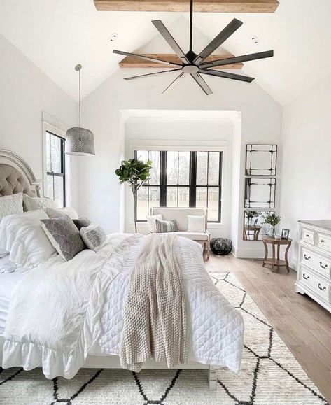 A modern bedroom is designed with a vaulted ceiling and rustic wood crossbeams over hardwood floors. Black framed windows illuminate the bed and white and black carpet, with an industrial-style pendant light hanging above the bedside table. Vaulted Ceiling Bedroom, Warm Wood Flooring, White Coverlet, Vaulted Ceiling Living Room, Ruffle Duvet Cover, Ball Chandelier, Modern Kitchen Island, White Comforter, White Carpet