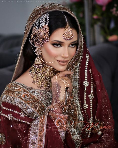 A stunning bride dressed in a classic red bridaL lehenga @prive___lux richly embroidered with intricate gold patterns. Her hair is styled in a traditional updo adorned with ornate jewelry, including a maang tikka, jhumkas, and a heavy necklace From @nk_collection . Her makeup features bold, expressive eyes with dramatic eyeliner and long lashes, complemented by a soft, glowing base. The highlight of her look is her nude lip color, adding a modern touch to the traditional ensemble. The backgro... Traditional Updo, Red Bridal Lehenga, Bridal Hair Up, Dramatic Eyeliner, Makeup Features, Anand Karaj, Heavy Necklace, Asian Bridal Makeup, Ornate Jewelry