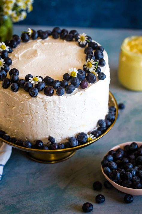 blueberry lemon curd cake on gold cake stand with bowl of blueberries in front and jar of curd in back. Blueberry Lemon Custard Cake, Blueberry Lemon Curd Cake, Lemon Blueberry Birthday Cake, Lemon Blueberry Wedding Cake, Lemon Blueberry Cake Decoration, Lemon Cake Decoration, Light Birthday Cake, Lemon Custard Cake, Bowl Of Blueberries