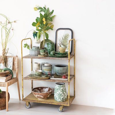 This metal vintage reproduction 3-tier cart on wheels is a stylish and practical piece of furniture that will enhance any room. It is crafted from high-quality metal that has a stunning distressed gold finish that adds charm and character to the cart. The cart has three rectangular shelves that offer ample space for storing and displaying items, use it as a bar cart, a kitchen cart, a bookcase, or a plant stand. The shelves have raised edges and handles that make them easy to access and secure. Bar Cart Essentials, Metal Cart, Metal Bar Cart, Old Warehouse, Household Plants, Organization Essentials, Rolling Cart, Color Making, Bygone Era