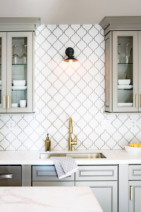 We loved how the arabesque pattern fits perfectly between the gray cabinets. Golden details and marble countertop give a luxurious feeling to the kitchen. The little small black lighting on the top of the faucet is a little cute touch that makes space a cozy place! Golden Backsplash Kitchen, White And Black Kitchen Backsplash Ideas, Gray Cabinet Backsplash Ideas, Kitchen Backsplash Dark Grout, White Tile Dark Grout Kitchen, White Backsplash Dark Grout, Light Tile Dark Grout, White Tile Black Grout Kitchen, Arabesque Backsplash Kitchen