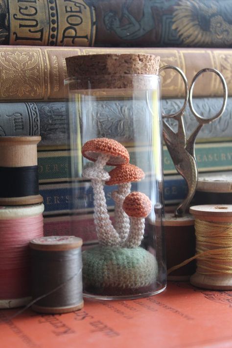 A collection of items placed in front of a stack of vintage books, including spools of thread, a pair of vintage bird scissors, and a glass jar filled with three orange capped micro crochet mushrooms with white stems sitting on a green mound of moss. Stash Jar Crochet Pattern, Crochet Mushroom Jar, Dark Cottagecore Crochet, Goblincore Crochet Ideas, Crochet Home Goods, Crochet Terrarium, Cottagecore Amigurumi, Crochet Display Ideas, Green Crochet Projects