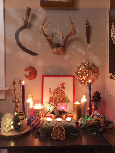 A small witch altar on a coffee table with antlers, antique scythe, antique knife, sun, moon, and a Christmas Santa cross stitch painting on the wall. The table has lot of lit winter holiday candles, a mini burning decorated Yule log, polar bear statue, chalice, deer statue candle holder, Mini light operated Christmas tree, decorated Yule besom with fake evergreen leaves berries and deer ornament, and an iron cauldron with a smudge stick and Palo santo incense stick. Yule Aesthetic Pagan Wallpaper, Winter Altar, Yule Altar, Yule Wicca Ritual, Lunar Witch Altar, Yule Mini Altar, Yule Pentacle, Witch Room, Wiccan Altar