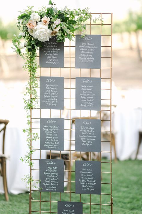 Gold grid escort card stand with greenery and flowers on the side and dark drey paper with table names and numbers in white calligraphy, film by Cavin Elizabeth Photography for a Gainey Vineyards Wedding Seating Chart Wedding Diy, Expo Ideas, Lemon Wedding, Bridal Expo, Vine Wedding, Tuscan Wedding, Santa Ynez, Seating Plan Wedding, Seating Cards