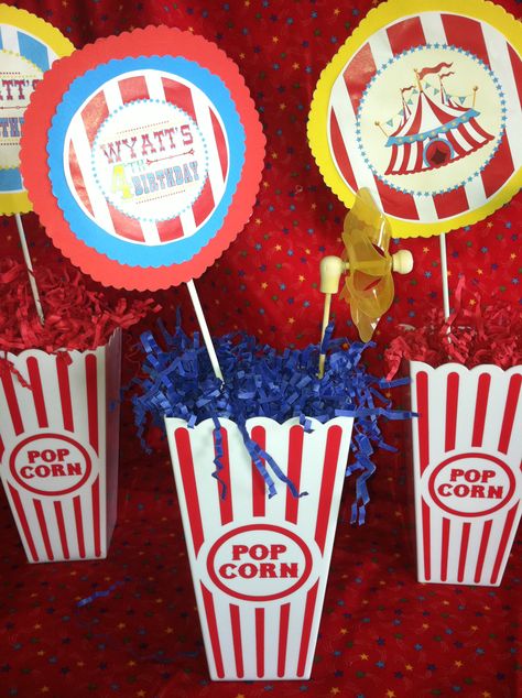 Table centerpieces.  Bought plastic popcorn containers at local dollar store.  Then I added rocks at the bottom (for weight) and red, yellow and blue shredded paper. The graphics are also by Metro Events.  I added the cardstock behind it then glued to a craft stick. I added pinwheels to some for color. Plastic Popcorn Containers, Popcorn Containers, Reception Table Centerpieces, Infant Room, Corn Pops, Backyard Reception, Popcorn Bucket, Shredded Paper, Carnival Party