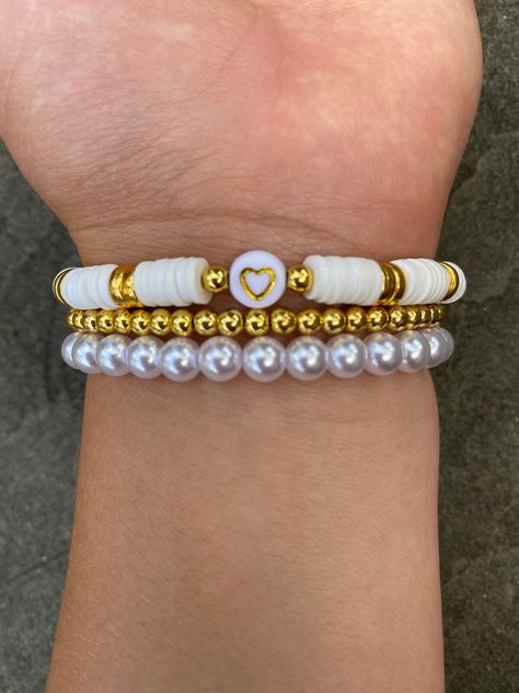 Stack of 3 bracelets, 4 mm gold beaded bracelet, 6 mm pearl bracelet, and a white clay bracelet with a gold heart. You may buy them as a set of three, or individually. Can be used for fancier occasions, or gifts. They are made with very stretchy and strong string that slides onto your wrist easily, therefore there is only one size. The tie on each bracelet is covered either by a pearl or a gold bead. Check out other items in my shop! Clay Bead Bracelet Ideas With Gold Beads, Matching Flat Bead Bracelets, Clay Bead With Pearls, White And Gold Clay Bead Bracelet, Fancy Clay Bead Bracelets, Bracelet Set Ideas, Preppy Bracelet Stack, Cool Bracelets, Make Clay Beads