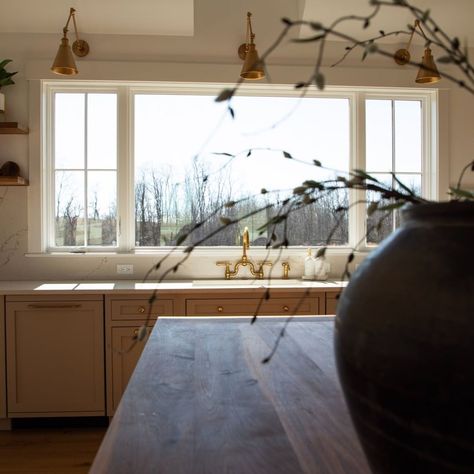 Three brass swing arm sconces are mounted over large kitchen windows. The marbled slab backsplash and brass faucet create a modern look when paired with neutral shaker cabinets sharing the same brass hardware. Window grilles are used to form sections, giving the large window a more homey appearance. Kitchen Windows Instead Of Upper Cabinets, Kitchen Window Next To Range, Wide Window Kitchen, Kitchen Window Ledge Ideas, Sink With Window Kitchen, Low Kitchen Window Solution, Windows Above Sink In Kitchen, Oven Between Two Windows, Arch Kitchen Window