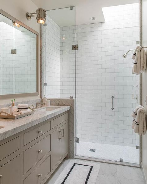 Sleek bathroom features a black framed white bathmat placed in front of a light taupe dual washstand fitted with satin nickel knobs and a gray marble countertop placed beneath a light taupe framed vanity mirror lit by a nickel industrial sconce mounted beside a seamless glass shower finished with white subway backsplash tiles and a polished nickel shower kit positioned under a skylight.