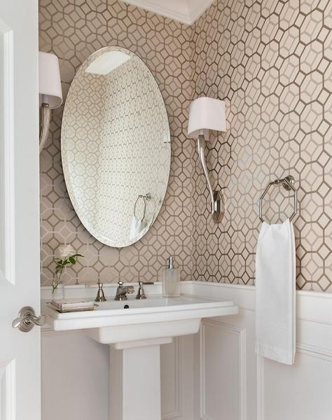 Flanked by Ruhlmann Single Sconces, an oval mirror, above a pedestal sink finished with a polished nickel faucet, is mounted to a wall accented with taupe hexagon wallpaper and lower wall wainscoting in this wonderfully designed white and taupe powder room. Half Bath Pedestal Sink, Wainscotting Wallpaper, Small Bathroom Update, Designer Bathroom Ideas, Spanish Beach House, Pedestal Sink Ideas, Powder Room Decor Ideas, Small Pedestal Sink, Floor Bathroom Ideas