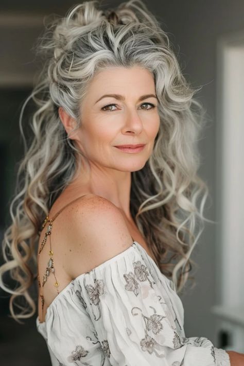 Elegant older woman with long, wavy gray hair and a white floral off-shoulder top, looking over her shoulder with a gentle smile. Long Curly Grey Hair Natural Curls, Long Curly Hair For Older Women, 2024 Summer Haircuts For Women, Long Curly Gray Hair Over 50 Curls, Gray Hair Updos Over 50, Over Fifty Hairstyles Over 50, Curly Silver Hair Over 50, Gray Hair Cuts Over 50, Silver Hair Updo