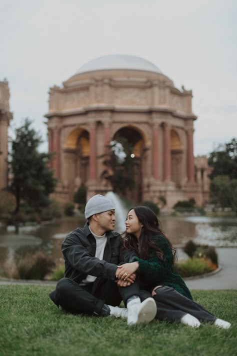 a man in a grey beanie sitting in the grass next to a brunette woman in a green fuzzy cardigan looking at each other and smiling with the dome and fountain of the palace of fine arts in san francisco in the background Golden Gate Park Photoshoot, Golden Gate Bridge Picture Ideas Couples, Palace Of Fine Arts San Francisco Engagement, San Francisco Couple Pictures, Palace Of Fine Arts Engagement Photos, Palace Of Fine Arts Photoshoot, Golden Gate Park Engagement Photos, Pink And Blue Sunset, San Francisco Engagement Photos