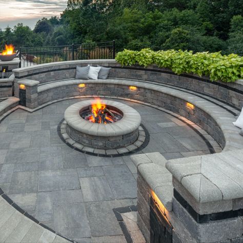 This family loves spending time together in their expansive outdoor entertaining space. An attractive curved staircase divides the space into two distinct areas: dining and entertaining on the upper level and poolside fun on the lower level.  Bristol Valley™ pavers were used throughout the space and Brussels Dimensional™ was used to create a wide variety of vertical features, from the garden and retaining walls to a complete outdoor kitchen, fire table, water features and a cozy firepit seating. Outdoor Fire Pit Area, Backyard Seating Area, Outdoor Fire Pit Designs, Patio Pavers Design, Outdoor Fireplace Designs, Fire Pit Landscaping, Sloped Backyard, Outdoor Fireplace Patio, Outdoor Patio Designs