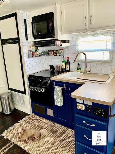 A picture of a renovated RV kitchen. It has white upper cabinets, blue lower cabinets, and a stick on subway tile backsplash. The floor has been redone and stained, and the refrigerator is painted white also. Navy Blue Camper Interior, Blue And White Rv Interior, Navy Blue Rv Interior, Camper Cabinet Removal, Blue Camper Cabinets, Blue Rv Interior, Navy Blue Kitchen Cabinets, Navy Blue Kitchen, Rv Exterior