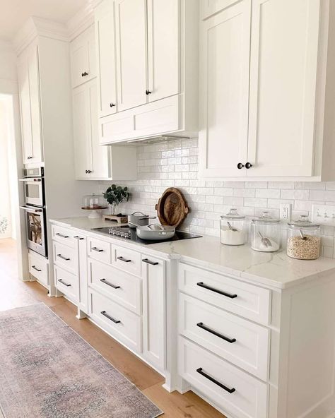 This kitchen has white upper and lower cabinets with a white subway tile backsplash. The countertops are white quartz and black handles and pulls are on the cabinets and drawers. The floor is hardwood with a faded rug. White Kitchen With Black Hardware, Kitchen With Black Hardware, Black Kitchen Handles, White Kitchen Countertops, White Shaker Kitchen Cabinets, Hardware Ideas, Off White Kitchens, Textured Rug, White Shaker Kitchen