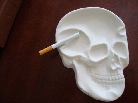 a cigarette sits along the side of a white ashtray. The ashtray is made out of clay, and is shaped like a 2-dimensional human skull. Air Dry Clay Ashtray, Skull Ashtray, Diy Skeleton, Skull Wedding Ring, Skull Engagement Ring, Kitchen Counter Top, Skull Wedding, Skeleton Skull, Ceramic Bottle