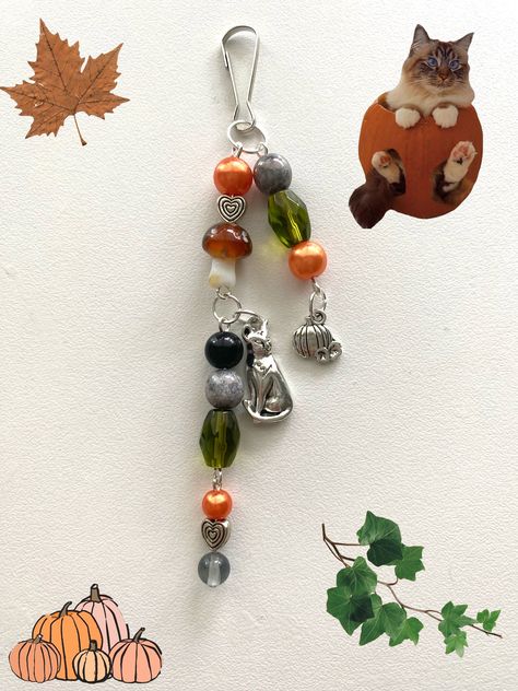 A beaded keychain with two silver charms. The pumpkin and cat charms along with the orange, green, and gray beads give off the fall aesthetic. The keychain also includes a glass mushroom bead, adding to the overall fall appearance. Orange Charm Bracelet, Fall Keychain Ideas, Keychain Ideas Diy Beads, Halloween Beaded Keychains, Beaded Keychains Aesthetic, Diy Charms Keychain, Keychain Beads Ideas, Fall Keychains, Beaded Keychain Ideas