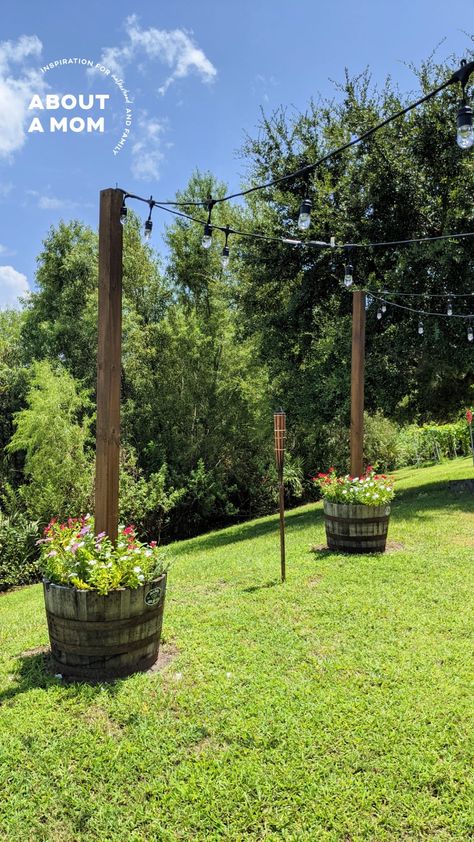 DIY Planter with Pole for String Lights - About a Mom Wood Poles For Outdoor String Lights, Front Yard String Lights, Acreage Yard Ideas, Wine Barrel Planter With Lights, Planters With Poles For Lights, Diy Light Pole Planter, Pole For String Lights, Wine Barrel Lighting, Poles For Outdoor Lights