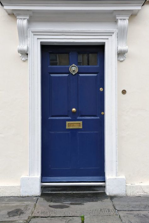 The main entrance has a white concrete facade. It has a blue door with a shiny brass knob right in the middle of it. The door also has a sculpted lion brass door knocker, lock, doorbell, and mail slot. The top part of the front door has glass panels. The door has a white brick molding. Farrow And Ball Hague Blue Front Door, Hague Blue Front Door Farrow Ball, Midnight Blue Front Door, Navy Blue Door Exterior, Hague Blue Exterior, Blue Exterior Door, Farrow And Ball Front Door Colours, Hague Blue Living Room, Dark Blue Front Door