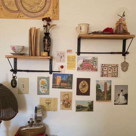 Two in uneven shelves with miscellaneous objects like teacups, books, and flowers. Beneath the shelf are postcards placed on the wall; different shape and space between they spread the wall with vibrant locations, floral drawings, and famous paintings. At the bottom of the picture is a small lamp with a rounded mirror made from a vintage cars side mirror. Maximalist Wall Shelves, Gallery Wall Postcards, Postcards Aesthetic Wall, Postcards Wall Decor, Wall Trinkets, Maximalist Shelf Decor, Postcards On Wall, Postcard Display Wall, Vintage Postcard Display