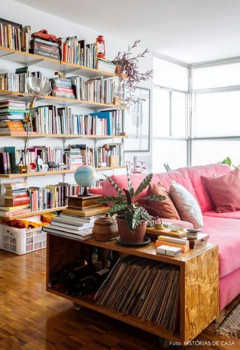 There is a rectangular oak end table in the foreground, with books on the upper and lower shelves and potted plants on top. Next to it there is a cotton candy pink couch with a few white and blush throw pillows. Behind the couch there is a large white wall with five long natural wood bookshelves. There are hundreds of books stacked on top of and next to each other on these shelves. There is a large window in the back window letting in tons of natural light. Colorful Eclectic Living Room, Behind Couch, Room Size Rugs, Boho Interior Design, Eclectic Living Room, Boho Interior, Furniture Arrangement, Small Living Room, Small Living