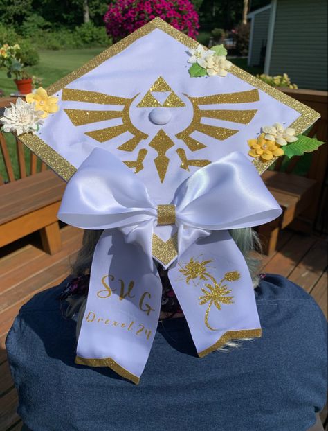 A white graduation cap with a sparkly gold boarder. The hat has yellow and white flowers as well as a sparkling gold triforce symbol. There is a white bow fixed to the bottom corner with the initials SVG  and the words “Drexel 24” on one tail of the bow. The other tail has an image of a silent princess in the same sparkling gold as the rest of the hats detail. Splatoon Graduation Cap, Grad Cap Studio Ghibli, Jjba Graduation Cap, Gojo Graduation Cap, Genshin Graduation Cap, Graduation Cap Designs Coraline, Legend Of Zelda Graduation Cap, Video Game Grad Cap, Undertale Graduation Cap