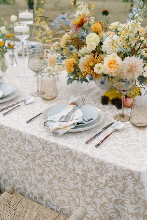 A wildflower floral arrangement and colorful wine glasses atop a neutral patterned linen with wicker chairs and a colorful napkin. Stradley Davidson's Ascot Garden was used for the napkin and Fitz Almond for the table linen. Colorful Wedding Linens, Patterned Wedding Table Linens, Patterned Tablecloth Wedding, Wildflower Table Setting, Floral Tablecloth Wedding, Wedding Drinkware, Vintage Table Setting, Reception Table Settings, Tying Knots