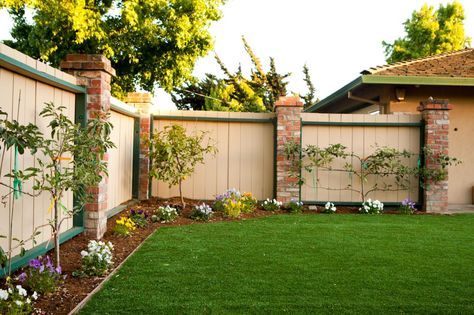 A border of small trees and beautiful flowers lines this well-maintained backyard. A beige fence with green trim and brick columns surrounds the space, providing privacy. Country Backyards, Fence Backyard, Landscaping Along Fence, Backyard Remodel, Diy Backyard Landscaping, Fence Landscaping, Ideas Backyard, Landscape Plans, Backyard Fences