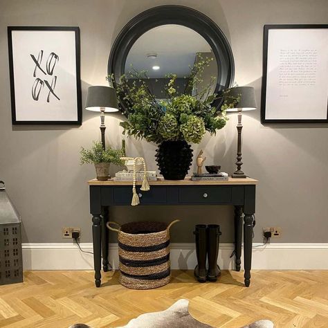 A two-toned black console table is styled with green, black and tan decor in this sophisticated gray entryway. The table is illuminated by gray turned wood entryway table lamps with gray shades. A round dark gray mirror is mounted above the table flanked by black frame artwork. Mirror Above Console Table, Vivienne Cutler, Round Mirror Decor Ideas, Black Console Table Decor, Gray Entryway, Tan Decor, Luxury Entryway, Foyer Makeover, Black Entryway