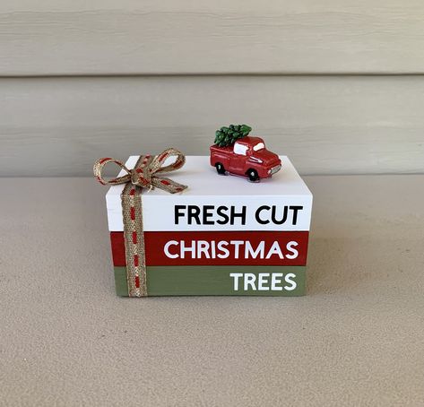 "This handmade mini wooden book stack features 3 rectangle blocks. The blocks are painted white, red, and green with permanent vinyl. The blocks display the words \" FRESH CUT CHRISTMAS TREES \" The blocks are glued together and has a red truck with Christmas tree that is also glued to the top and has a ribbon tied into a bow.  The book stack is about 3\"W x 4\"L x 2.5\" H These will ship in 3 days from ordering Thanks for shopping at  Farmhouse Charm To Love" Wooden Book Decor, Mini Book Stacks Diy, Christmas Book Stacks, 2 X 4 Crafts, Red Truck With Christmas Tree, Holiday Luncheon, Tiered Tray Decor Christmas, Stack Books, Craft Signs