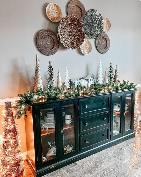 A black farmhouse sideboard with glass doors is set along the wall in this living room. A Christmas village with miniature trees and draping garland is set on top, along with small white lights. Round woven baskets are hung on the wall above, with two twig trees wrapped in white lights are set on either side. Christmas Sideboard Decor, Buffet Cabinet Decor, Buffet With Glass Doors, Taupe Dining Room, Decorating Sideboards, Decor With Baskets, Sideboard With Glass Doors, Modern Farmhouse Sideboard, Modern Buffet Table