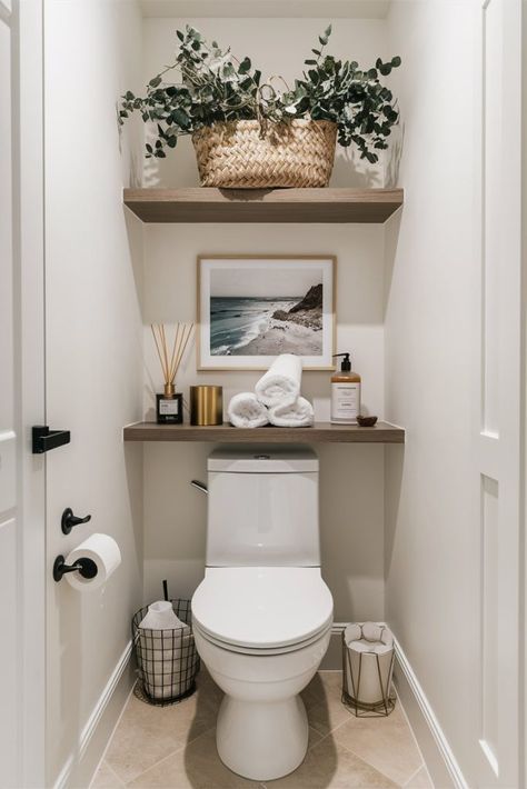 A bathroom with over-the-toilet decor featuring two wooden shelves. The top shelf holds a large woven basket filled with green plants. The lower shelf displays rolled white towels, a scented diffuser, a soap dispenser, and a coastal photograph framed in white. The toilet area also includes a wire basket for toilet paper and a gold wastebasket. The walls are white, creating a clean and serene atmosphere. Small Half Bathroom, Half Bathroom Decor, Toilet Room Decor, Bathroom Counter Decor, Bathroom Shelf Decor, Restroom Decor, Bathroom Decor Apartment, Toilet Room, Bathroom Design Decor