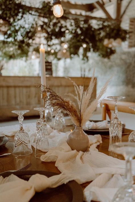 This shows a close up of a rustic barn dining tablescape. The focus is on a wooden table with white table runners on it, gold  candlesticks with LED candles, cream bud vases with dried flowers, cutlery and tableware. Dried Flower Table Decor, Dried Flower Arrangements Wedding Table, Dried Flower Table Arrangements, Small Table Design, Dining Table Flowers, Barn Photography, Vase Small, Flower Installation, Wedding Arrangements