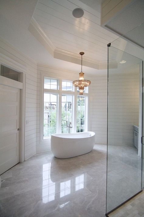 Stunning master bathroom features an octagon tray ceiling illuminated by an empire style chandelier suspended above a modern egg shaped tub and gooseneck tub filler situated in front of a three large windows framed by tongue and groove paneling atop gray stone tiled floors. Chandelier Over Tub, Grey Stone Tiles, Veranda Interiors, Porcelain Tub, Stone Tile Flooring, Bathroom Vanity Tray, Tongue And Groove Panelling, Painting Wood Paneling, Grey Bathroom Vanity