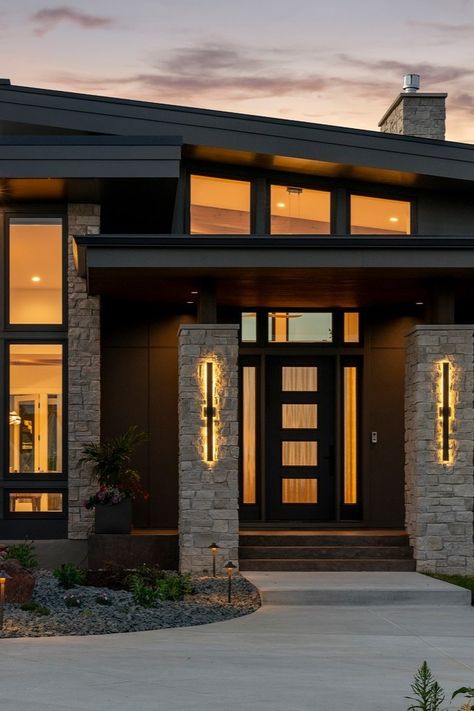 The stone exterior, complemented with bronze Andersen windows, not only adds visual interest but also lends a timeless quality to the home’s facade, fulfilling the homeowners’ desire for a luxurious yet durable exterior finish. ____ Front door, home exterior, lights on sunset shot photography, natural stone entrance, slanted roof, modern home design. Home Exterior Lights, Stone Entrance, Slanted Roof, Stone Exterior Houses, Glass Exterior, House Flip, Exterior Lights, Andersen Windows, Stone Exterior