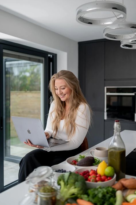 A woman with long blonde hair sits at a kitchen counter using a laptop. The counter is covered with various fruits, vegetables, and a bottle of olive oil. Large windows are seen in the background, creating an inspiring scene perfect for Liverpool brand photography or entrepreneur portraits by a UK branding photographer. Lifestyle Kitchen Photoshoot, Health Coaching Aesthetic, Dietitian Branding Photoshoot, Dietician Branding Photoshoot, Health And Wellness Coaching Business, Nutritionist Photoshoot Health Coach, Nutrition Coach Photography, Nutrition Coach Photoshoot, Health Wellness Photoshoot