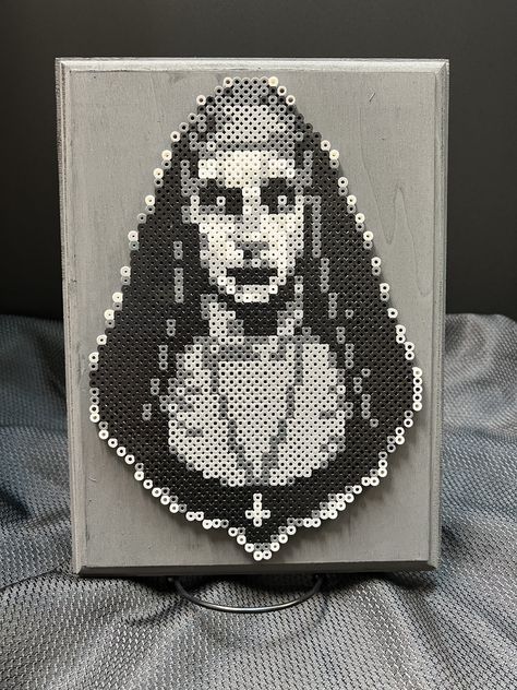 It's Valak the scary Nun from the popular Conjuring Universe horror films! Made from perler beads and mounted on a wood canvas, this creepy piece of art would look great hanging anywhere! Picture hanger included on back for easy hanging! Horror Movie Perler Bead Patterns, Fuse Beads Halloween, Hama Beads Horror, Wednesday Addams Perler Beads, Scream Perler Bead Patterns, Spooky Perler Bead Patterns, Scream Perler Beads, Scary Pixel Art, Horror Perler Beads