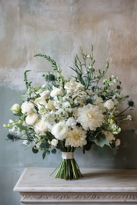 A large, elegant bouquet of white flowers including roses, dahlias, and ranunculus, accented with greenery and small blue thistles, arranged on a marble surface against a textured gray wall. Sage Green Flower Bouquet, White And Green Bridesmaid Bouquet, Wedding Bouquets Green, Green And White Winter Wedding, Wedding Flowers White And Green, May Wedding Flowers, Green And White Wedding Bouquet, Sage Green Wedding Flowers, White And Green Wedding Bouquet